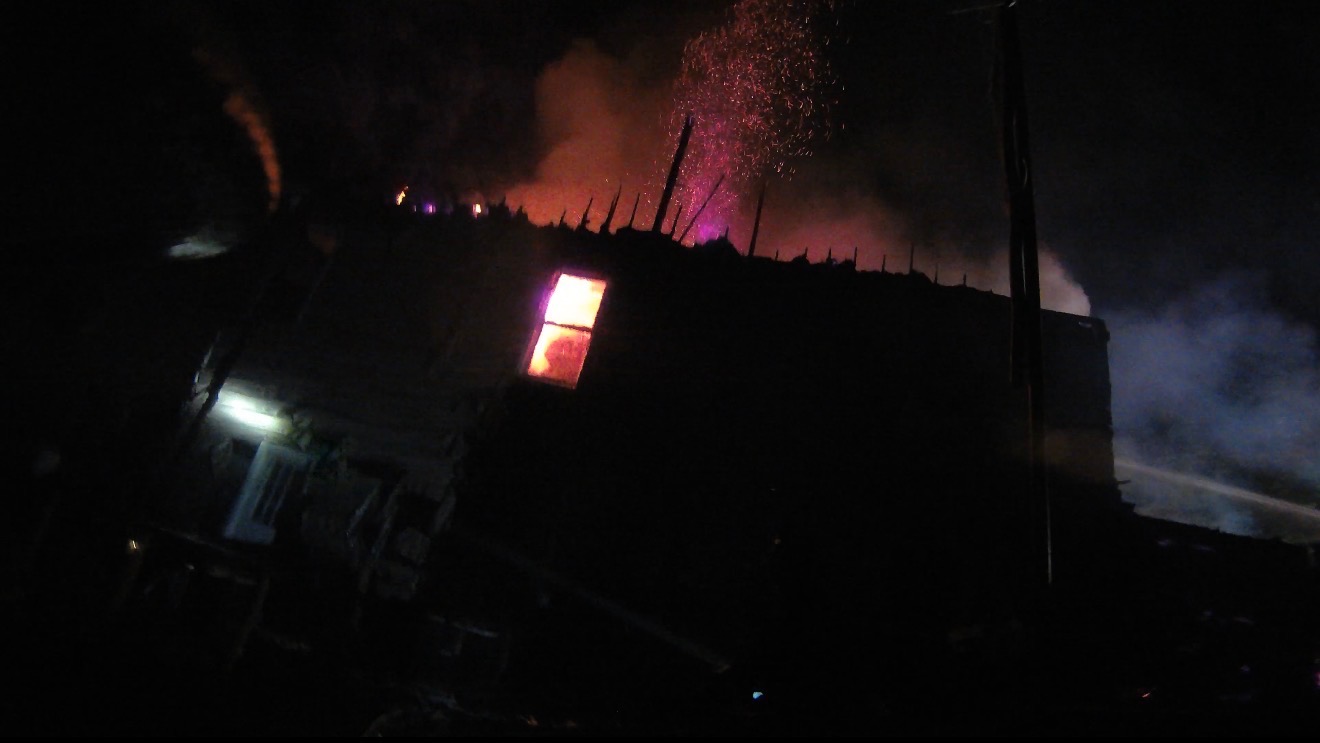 Fire billows from a burned out roofline of a residential home at night silhouetted by the night sky. 