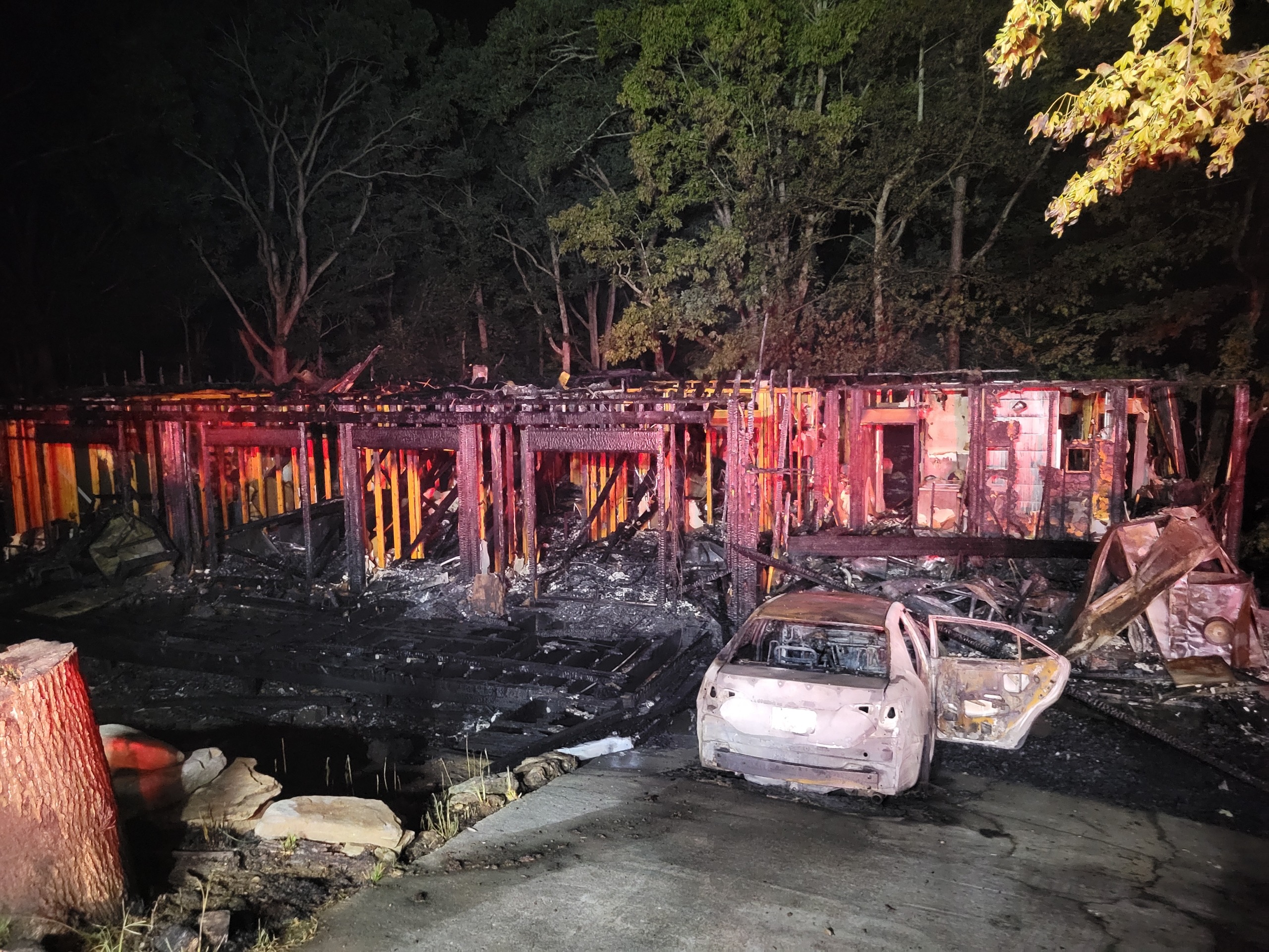 A heavily damaged home is shown after the fire has been extinguished. The structure is reduced to a charred frame, and a burned vehicle sits in the driveway surrounded by fire debris.